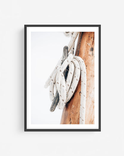 Fine art nautical rope photograph in a black frame showing a coiled rope wrapped around a metal cleat on a weathered wooden mast.