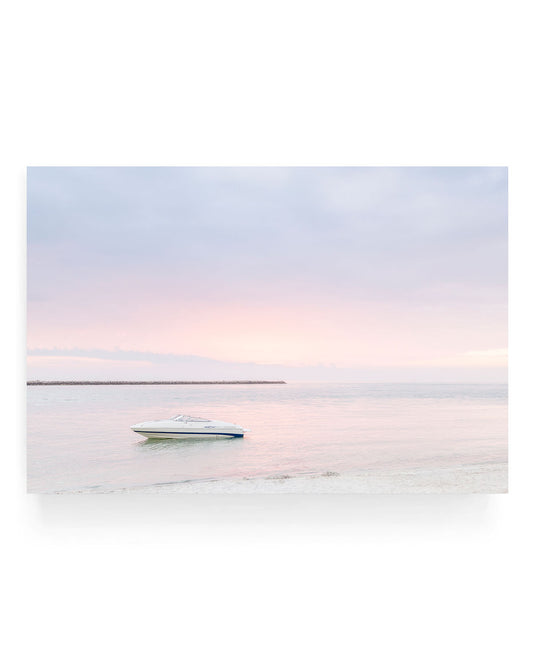 White speedboat floating on calm pastel ocean water at Clearwater Beach during soft pink and blue sunset, with a distant breakwater and smooth sandy shoreline in view.