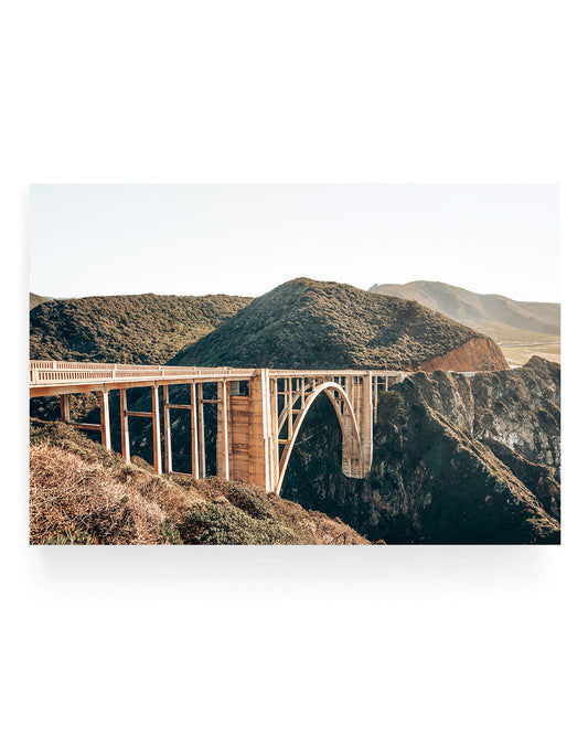 Golden hour photograph of Bixby Creek Bridge in Big Sur California, featuring the iconic concrete arch bridge spanning a rugged coastal canyon with rolling green hills and warm sunset light illuminating the landscape.