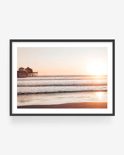 Huntington Beach sunset photography in black frame showing pier, waves, and golden light, coastal wall art print.