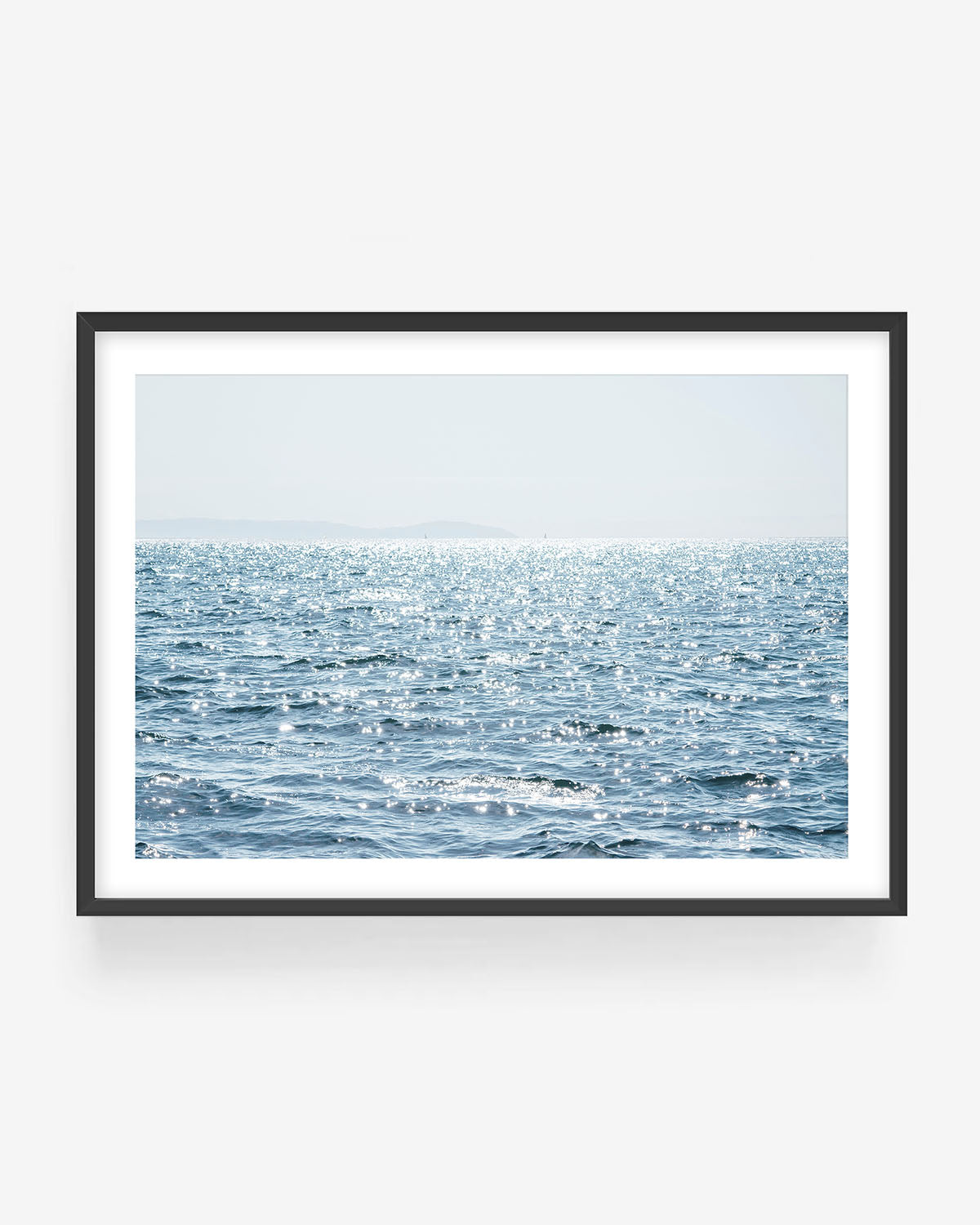 Minimal ocean wall art showing sunlit blue water and sparkling sea surface, displayed in a black frame, serene coastal photography print for modern minimalist home decor
