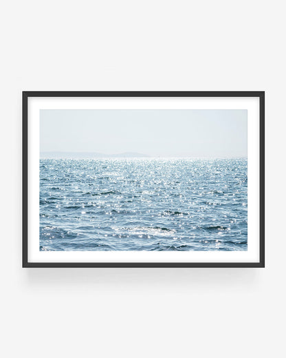 Minimal ocean wall art showing sunlit blue water and sparkling sea surface, displayed in a black frame, serene coastal photography print for modern minimalist home decor
