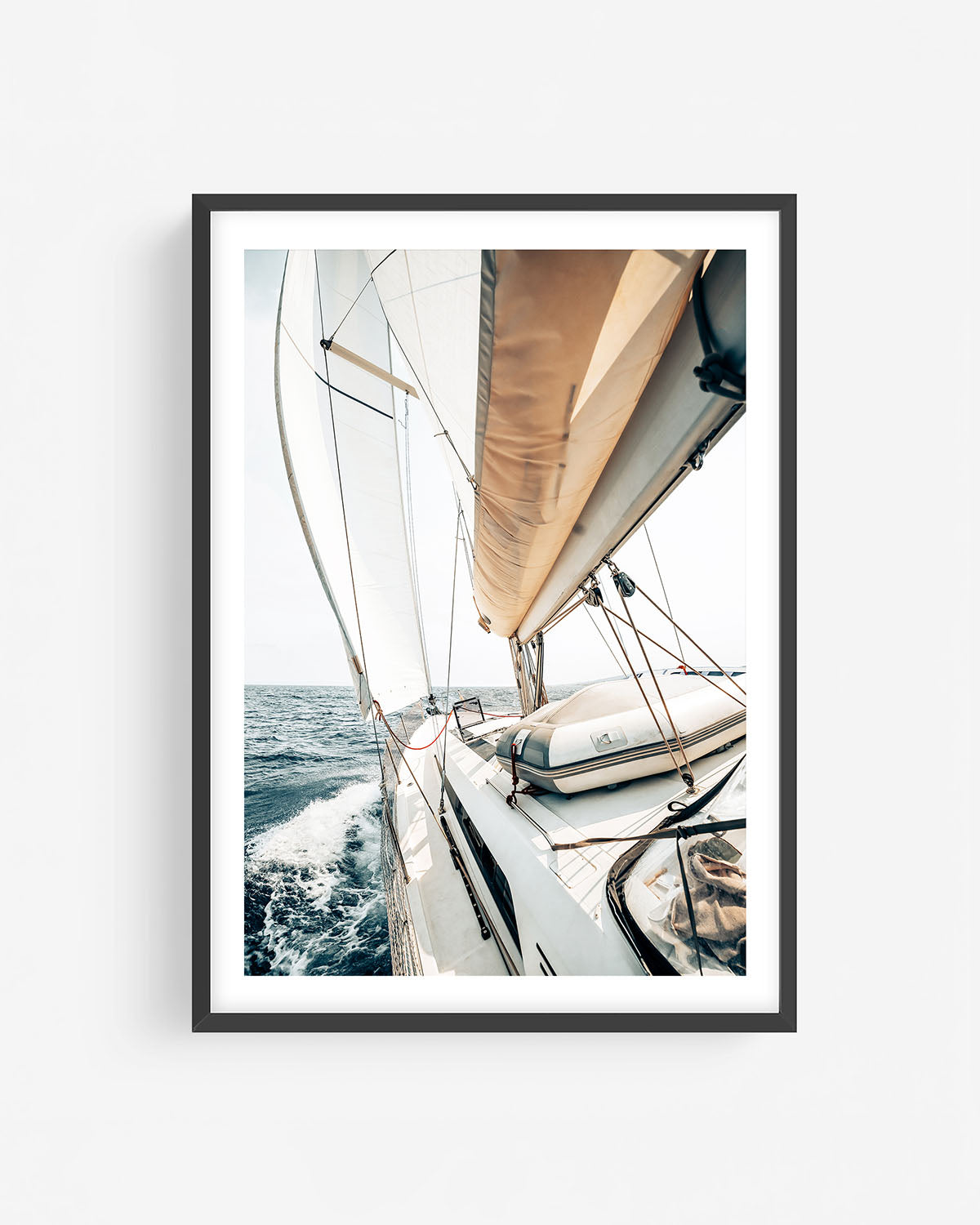 Fine art sailing photograph in a black frame showing a sailboat deck, sunlit sails, and open sea with dynamic water and coastal light.