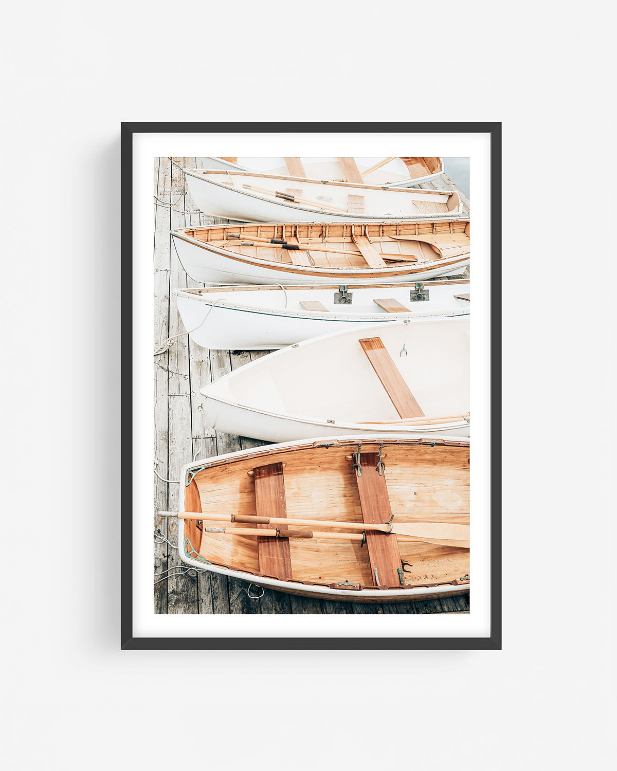 Fine art rowboat photograph in a black frame showing wooden and white boats stacked on a dock in a clean, coastal composition.