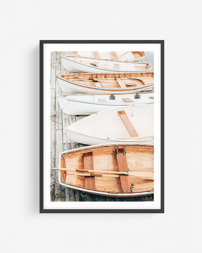 Fine art rowboat photograph in a black frame showing wooden and white boats stacked on a dock in a clean, coastal composition.