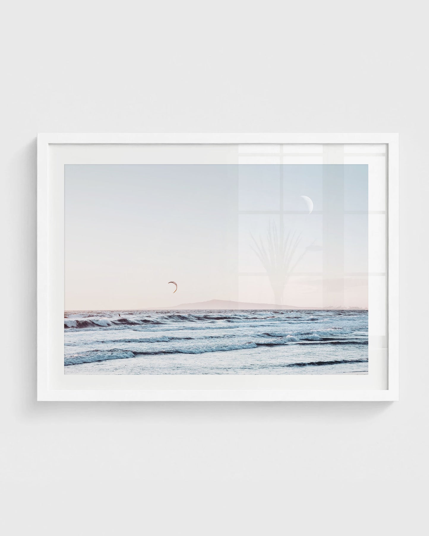 Huntington Beach wall art featuring ocean waves with a distant kite surfer, a soft pastel sky with a visible moon, and a faint horizon line, framed in a white frame.