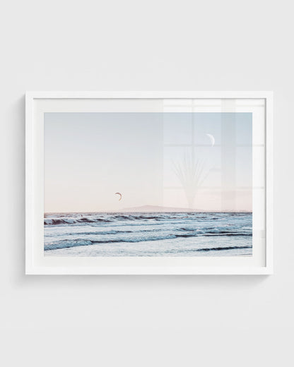 Huntington Beach wall art featuring ocean waves with a distant kite surfer, a soft pastel sky with a visible moon, and a faint horizon line, framed in a white frame.