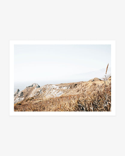 Big Sur landscape photography featuring sun-bleached grasses, rugged coastal bluffs and distant mountains fading into soft Pacific haze.