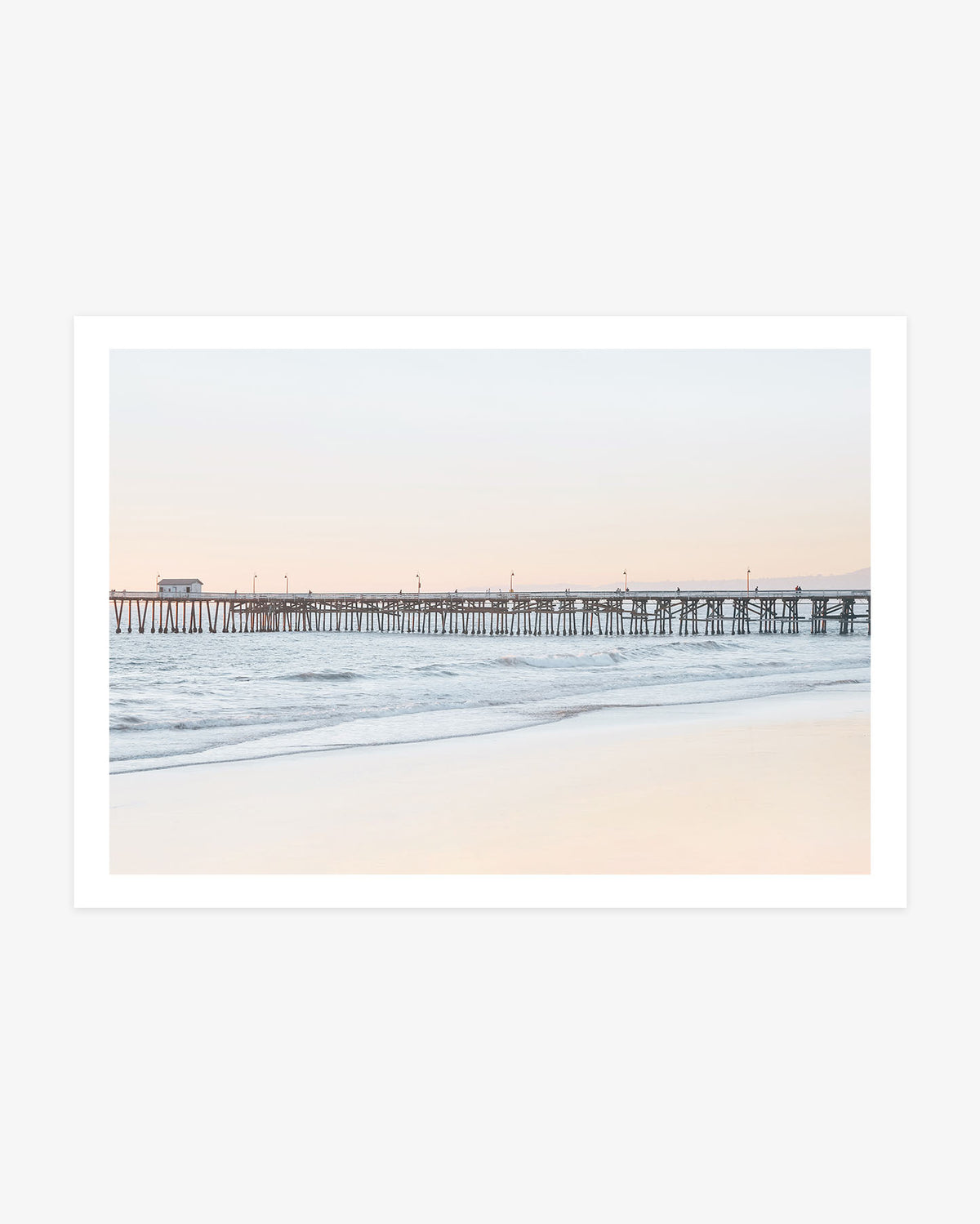 San Clemente Pier wall art featuring the long wooden pier above gentle Pacific waves beneath a pale blue and blush coastal sky.