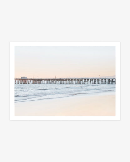 San Clemente Pier wall art featuring the long wooden pier above gentle Pacific waves beneath a pale blue and blush coastal sky.