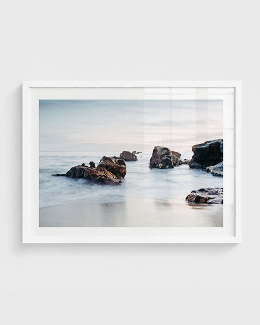 Laguna Beach wall art featuring rocky shoreline formations in calm ocean water with soft waves, wet sand, and a muted sky, framed in a white frame.