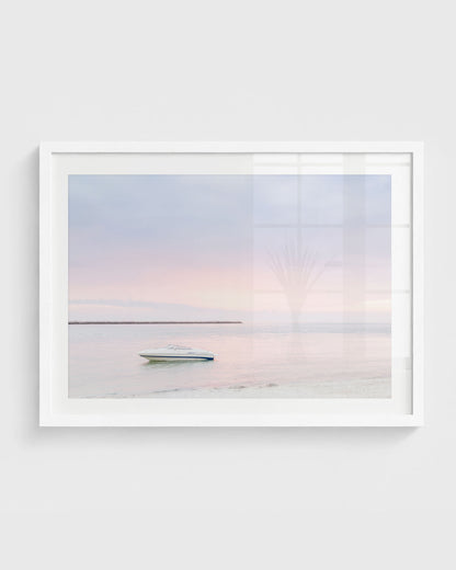 Clearwater Beach sunset wall art featuring a small white boat floating on calm water with a soft pastel pink and blue sky and a distant shoreline, framed in a white frame.