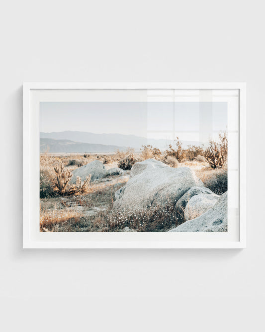 California desert wall art featuring large sunlit boulders, dry grasses, scattered cacti, and distant mountains in soft neutral tones, framed in a white frame.