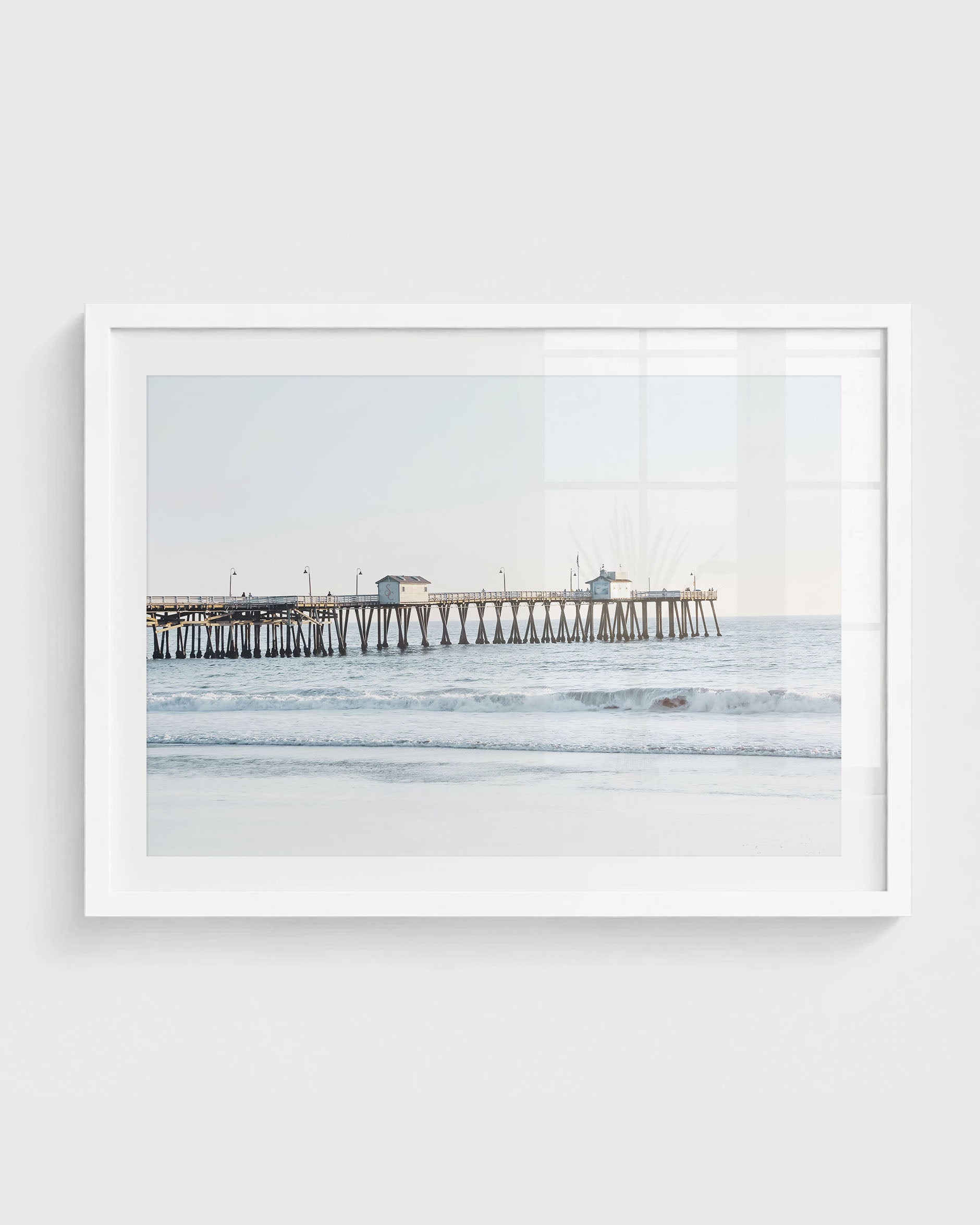 San Clemente Pier wall art featuring a long wooden pier extending over soft ocean waves with a sandy shoreline and a clear pastel sky, framed in a white frame.