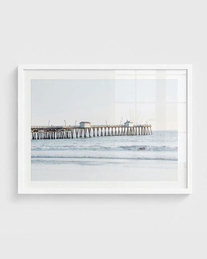San Clemente Pier wall art featuring a long wooden pier extending over soft ocean waves with a sandy shoreline and a clear pastel sky, framed in a white frame.
