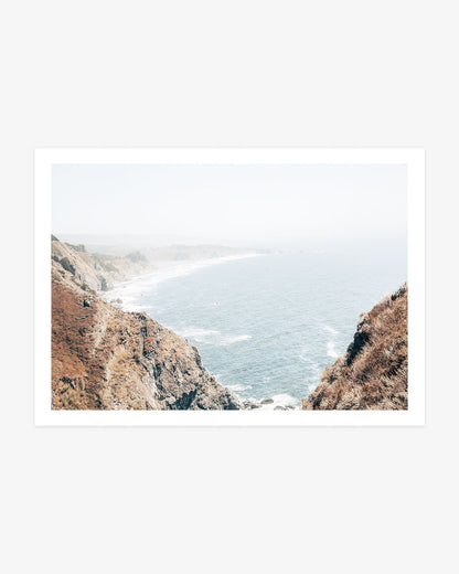 Big Sur coastal photography featuring steep sunlit cliffs framing a sweeping view of the blue Pacific and a hazy shoreline stretching into the distance.