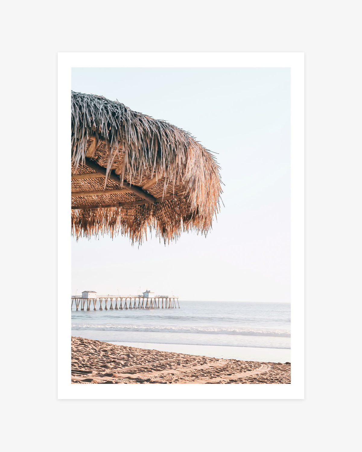 San Clemente beach wall art featuring a close view of a thatched palapa with the sandy shoreline, calm Pacific Ocean and pier beyond.