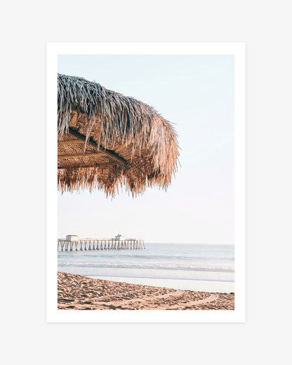 San Clemente beach wall art featuring a close view of a thatched palapa with the sandy shoreline, calm Pacific Ocean and pier beyond.