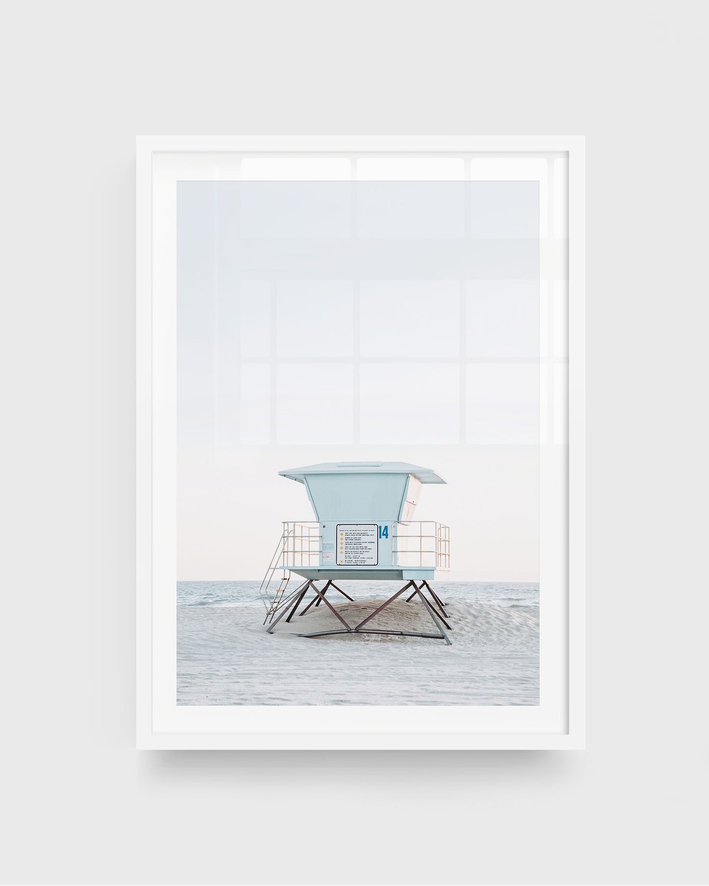 Huntington Beach lifeguard tower wall art featuring a centered pale blue lifeguard tower on stilts with railings, set on a sandy beach with soft ocean waves and a light sky, framed in a white frame.