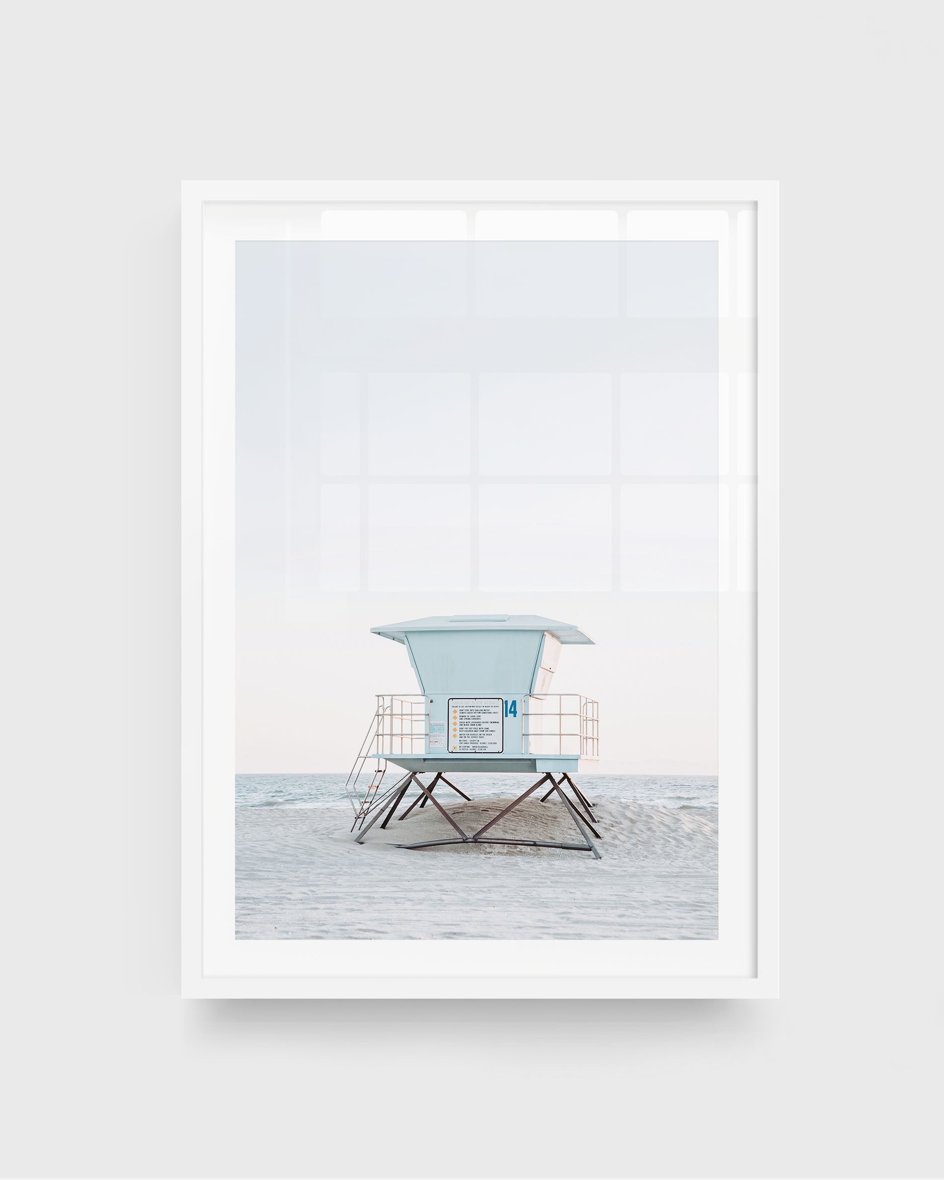 Huntington Beach lifeguard tower wall art featuring a centered pale blue lifeguard tower on stilts with railings, set on a sandy beach with soft ocean waves and a light sky, framed in a white frame.