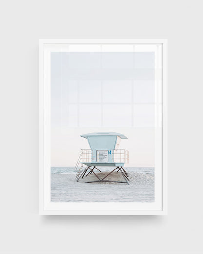 Huntington Beach lifeguard tower wall art featuring a centered pale blue lifeguard tower on stilts with railings, set on a sandy beach with soft ocean waves and a light sky, framed in a white frame.