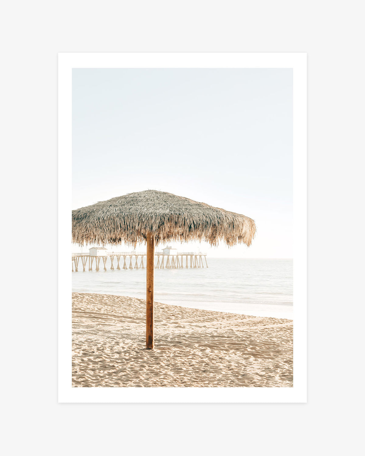 San Clemente beach umbrella wall art featuring a thatched palapa on the sand with the pier and calm Pacific Ocean in the background.