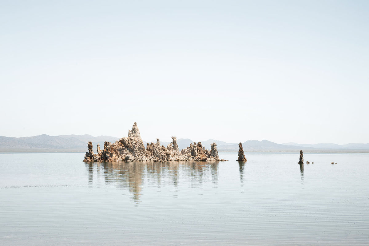 Minimalist Mono Lake landscape with tufa formations reflected in calm water beneath a pale blue sky and distant mountains.