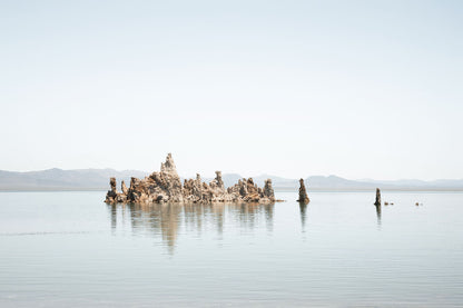 Minimalist Mono Lake landscape with tufa formations reflected in calm water beneath a pale blue sky and distant mountains.