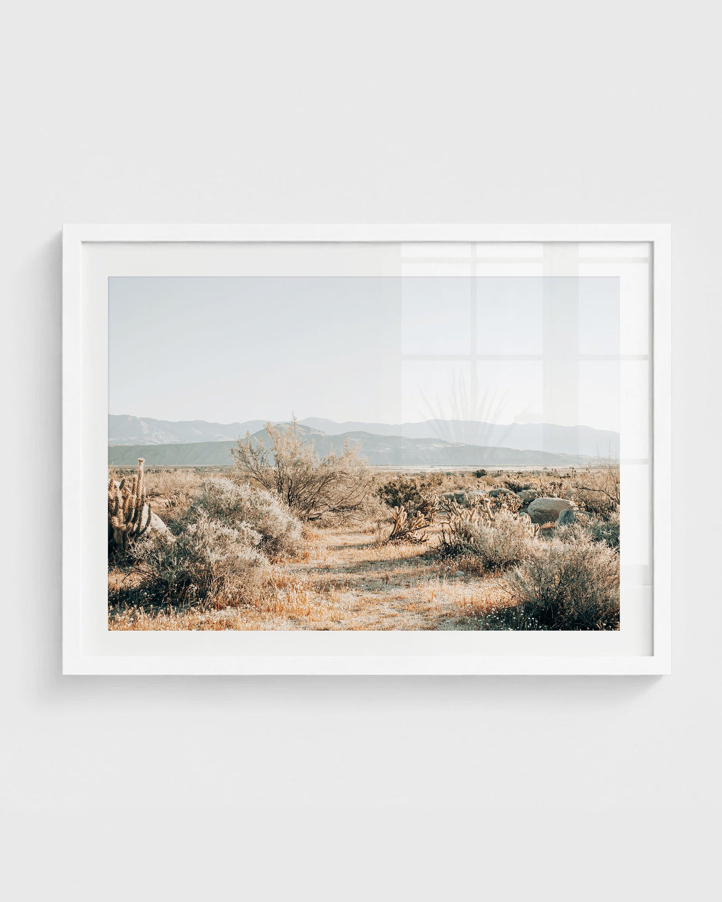 California desert wall art featuring a sandy path through dry grasses, scattered cacti and shrubs, with distant mountains under a soft clear sky, framed in a white frame.