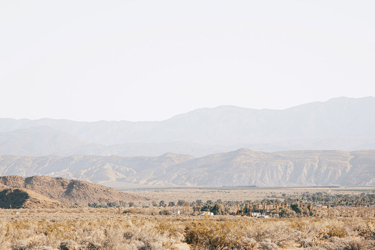 Expansive Anza-Borrego desert landscape with layered mountains, sandy terrain, and palms beneath a pale California sky.