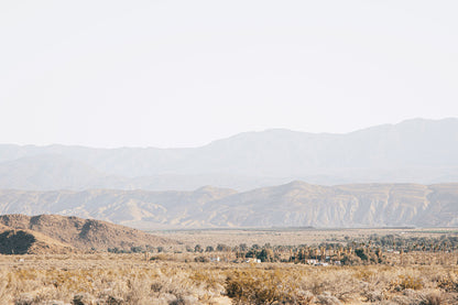 Expansive Anza-Borrego desert landscape with layered mountains, sandy terrain, and palms beneath a pale California sky.