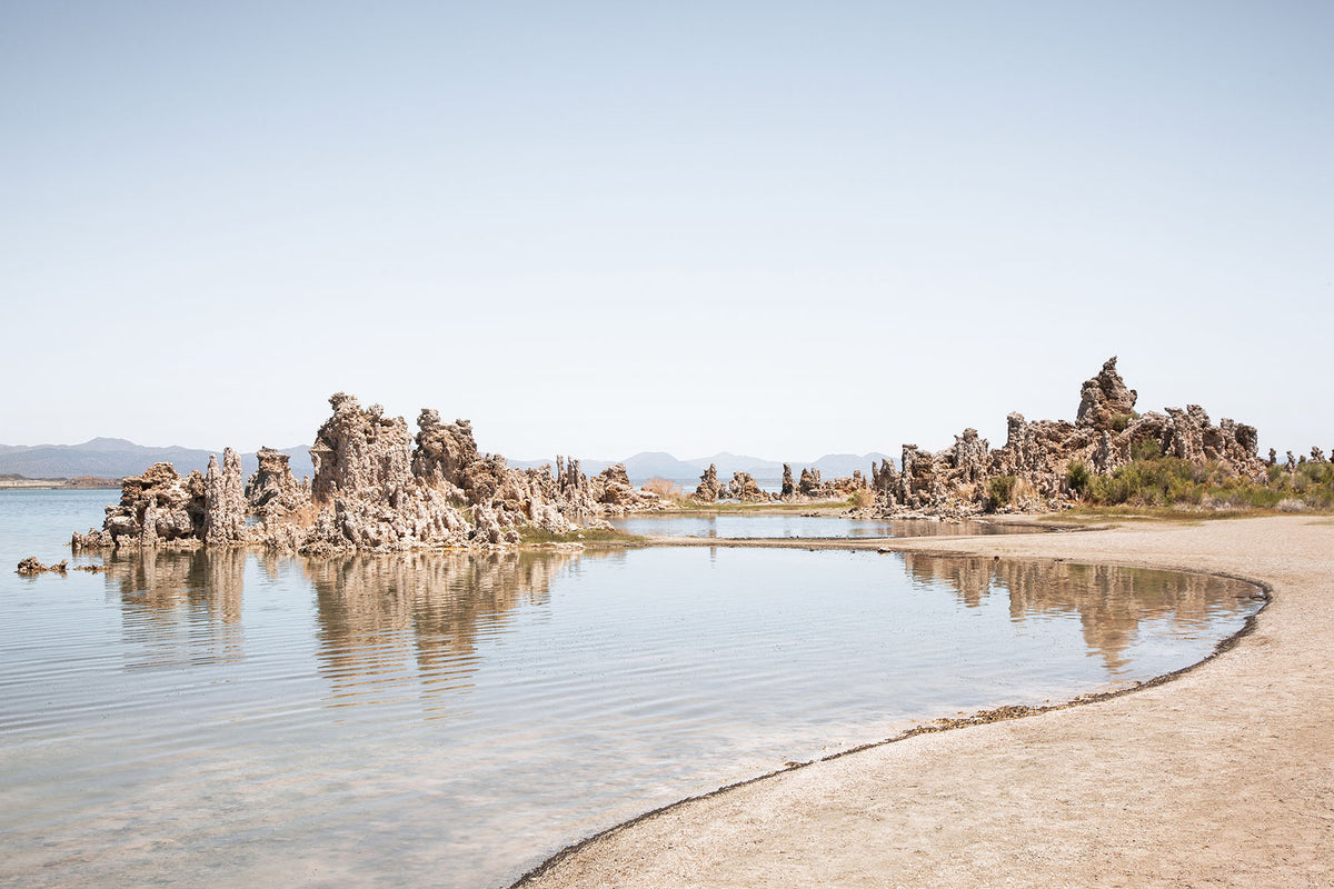 Mono Lake shoreline with sculptural tufa formations reflected in calm water beneath a pale blue California sky.
