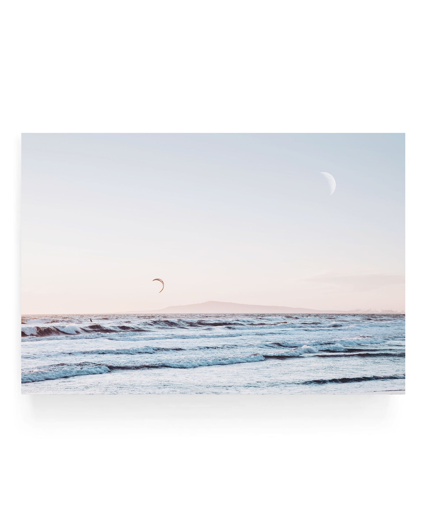 Ocean waves rolling toward shore under a soft pastel sky with a crescent moon above and a kite surfer in the distance, California coastal seascape photography with calm blue water and distant island horizon.