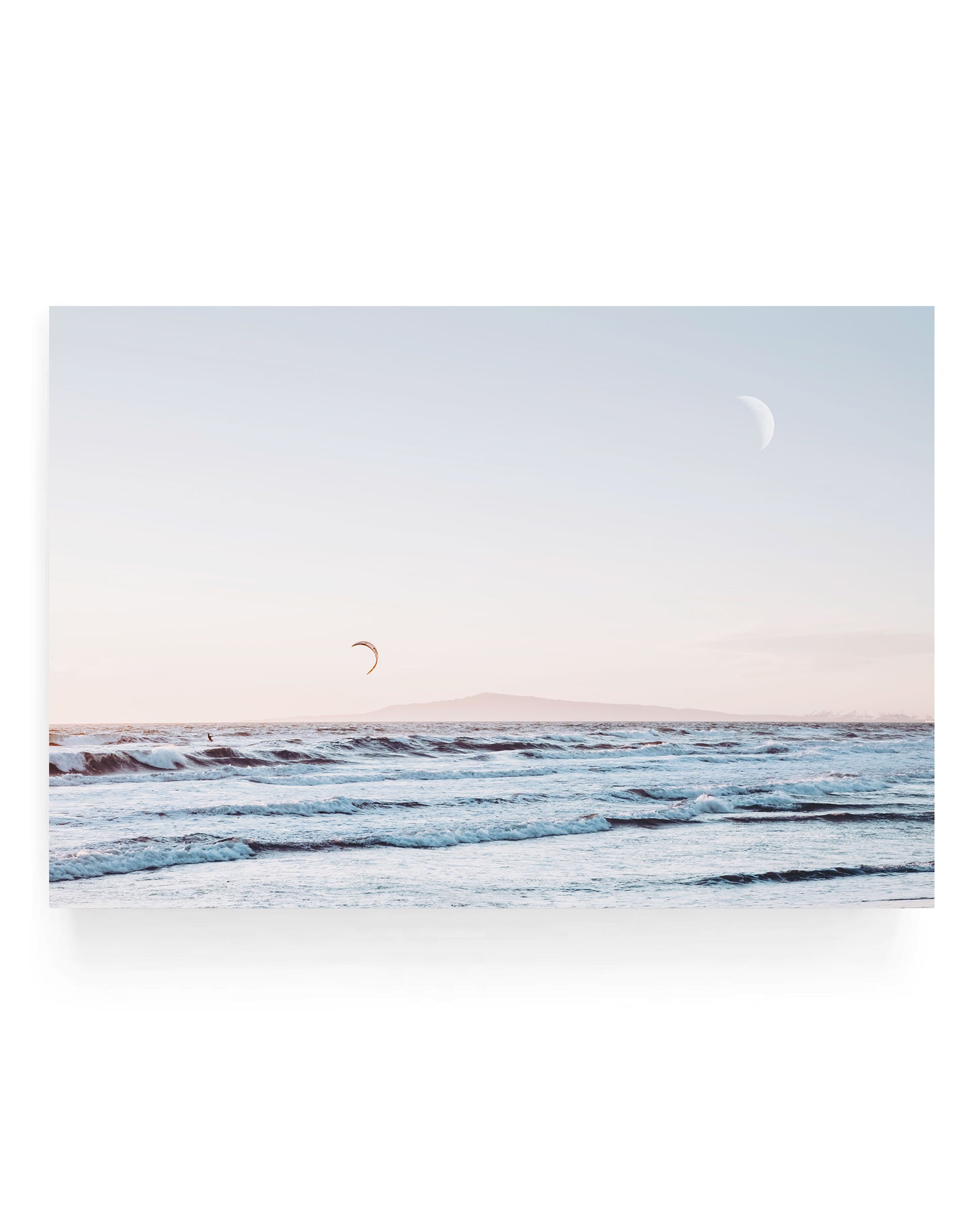 Ocean waves rolling toward shore under a soft pastel sky with a crescent moon above and a kite surfer in the distance, California coastal seascape photography with calm blue water and distant island horizon.