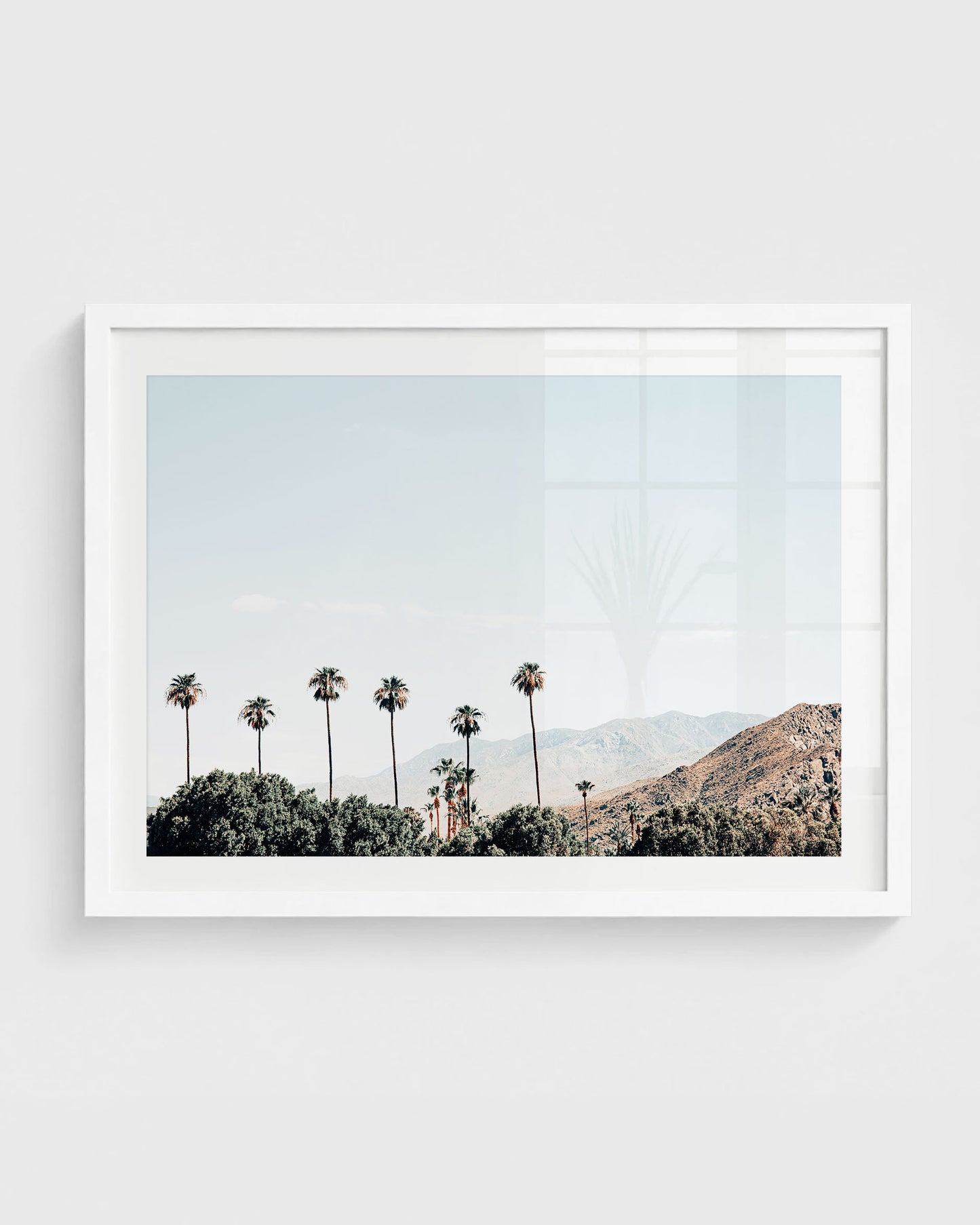 Palm Springs wall art featuring tall palm trees rising above desert greenery with mountain ranges in the background under a clear blue sky, framed in a white frame.