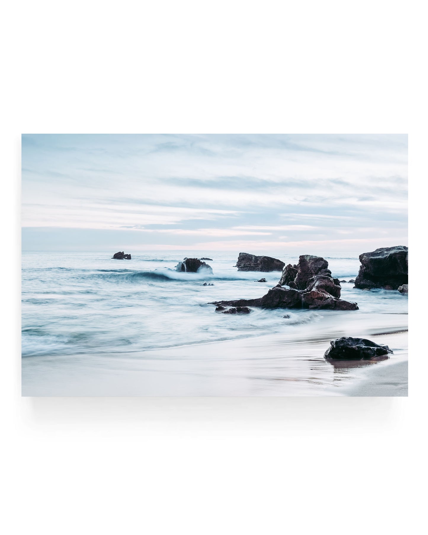 Laguna Beach rocky shoreline at sunset with soft ocean waves rolling toward sandy beach and dark coastal rocks under a pastel blue and pink sky, long exposure coastal landscape photography.