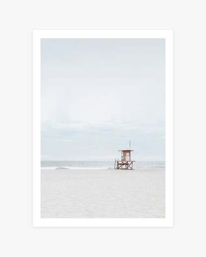 Newport Beach lifeguard tower wall art featuring Tower 65 standing on a wide sandy shore beneath a pale blue sky beside the Pacific Ocean.