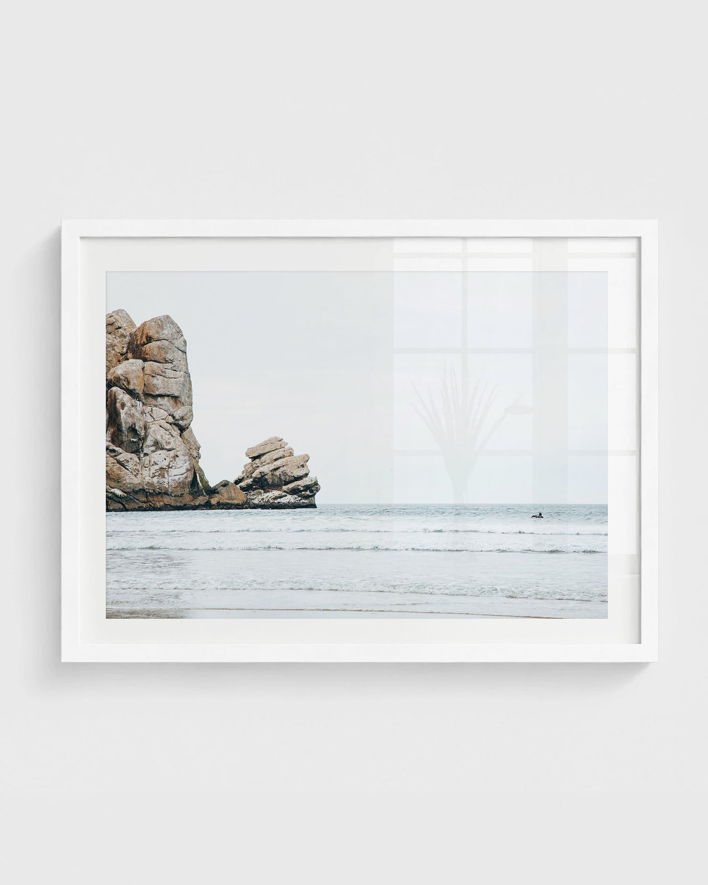 Morro Bay wall art featuring large coastal rock formations rising from the ocean with gentle waves and a distant surfer under a soft overcast sky, framed in a white frame.