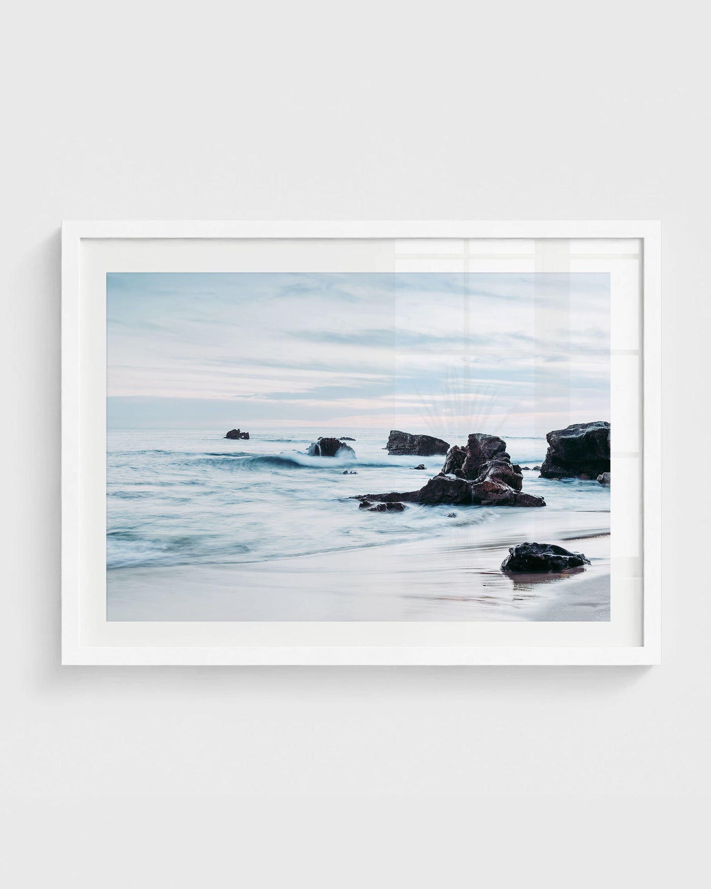Laguna Beach wall art featuring ocean waves rolling around dark rocky formations along the shoreline with wet sand and a soft cloudy sky, framed in a white frame.