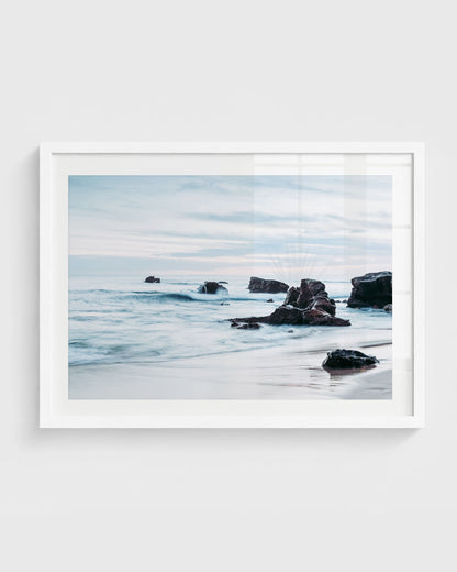 Laguna Beach wall art featuring ocean waves rolling around dark rocky formations along the shoreline with wet sand and a soft cloudy sky, framed in a white frame.