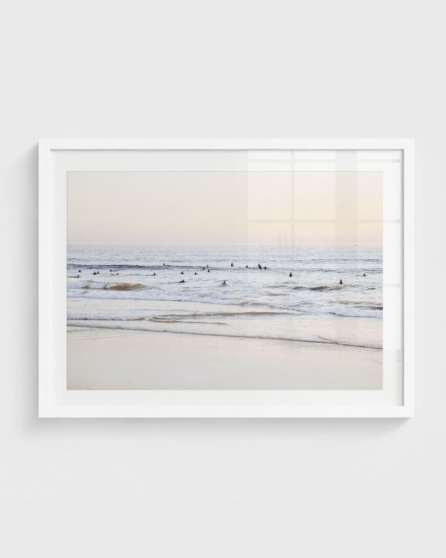 Beach surfers wall art featuring a group of surfers sitting in gentle ocean waves near the shoreline under a soft pastel sky, framed in a white frame.