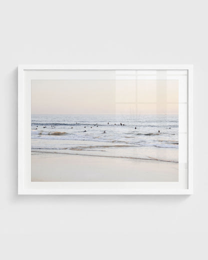 Beach surfers wall art featuring a group of surfers sitting in gentle ocean waves near the shoreline under a soft pastel sky, framed in a white frame.