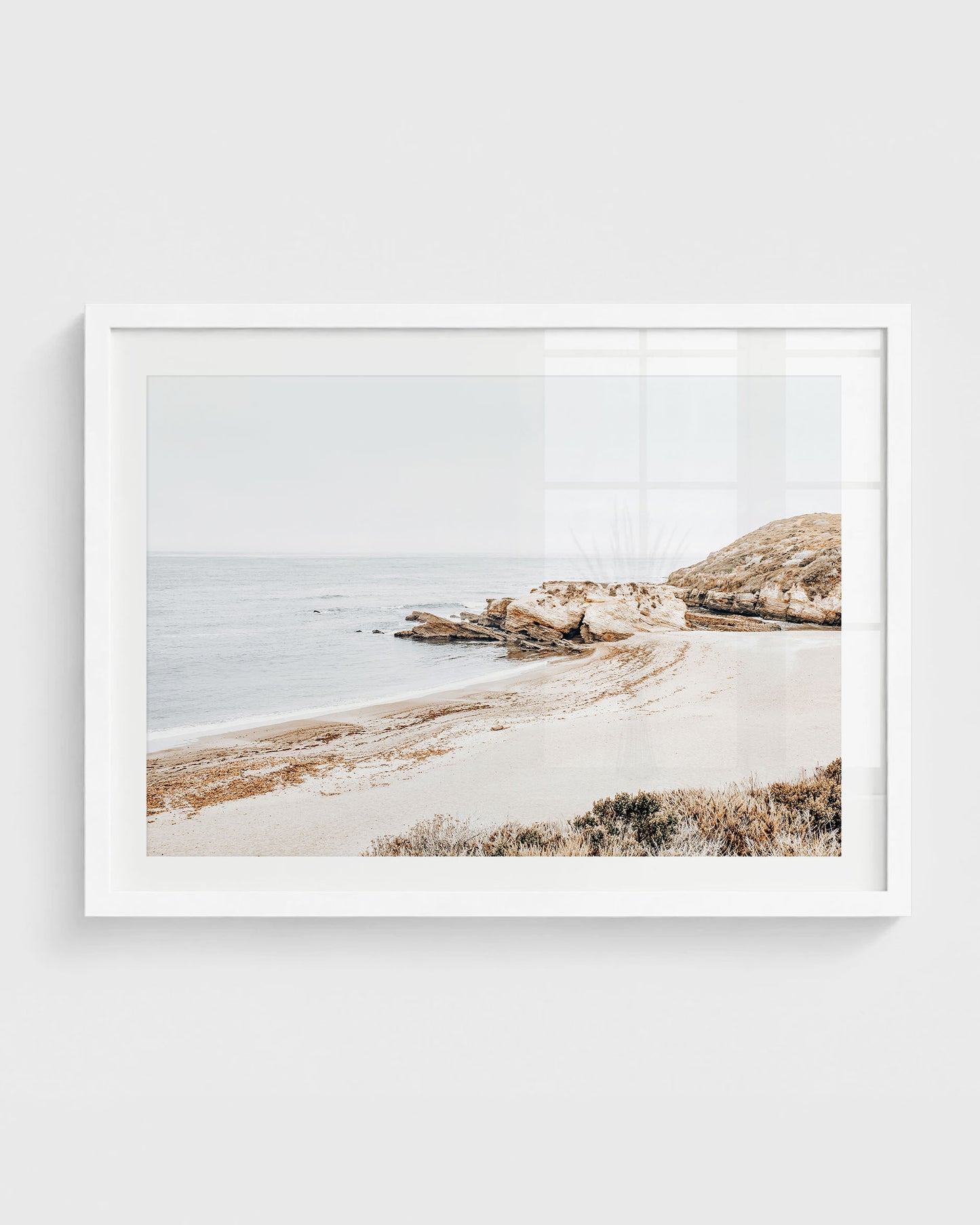 Montaña de Oro beach wall art featuring a quiet sandy cove with rocky cliffs, soft ocean water, and a muted overcast sky, framed in a white frame.