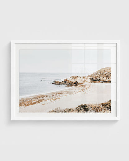 Montaña de Oro beach wall art featuring a quiet sandy cove with rocky cliffs, soft ocean water, and a muted overcast sky, framed in a white frame.