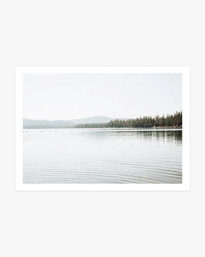 Northern California lake photography featuring calm reflective water, an evergreen shoreline and softly layered forested hills beneath a pale sky.