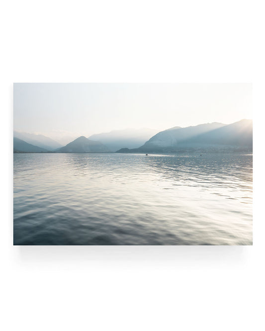 Serene Lake Lago Maggiore landscape wall art featuring calm reflective water, soft golden sunlight, hazy mountain silhouettes, and a peaceful shoreline in Northern Italy.