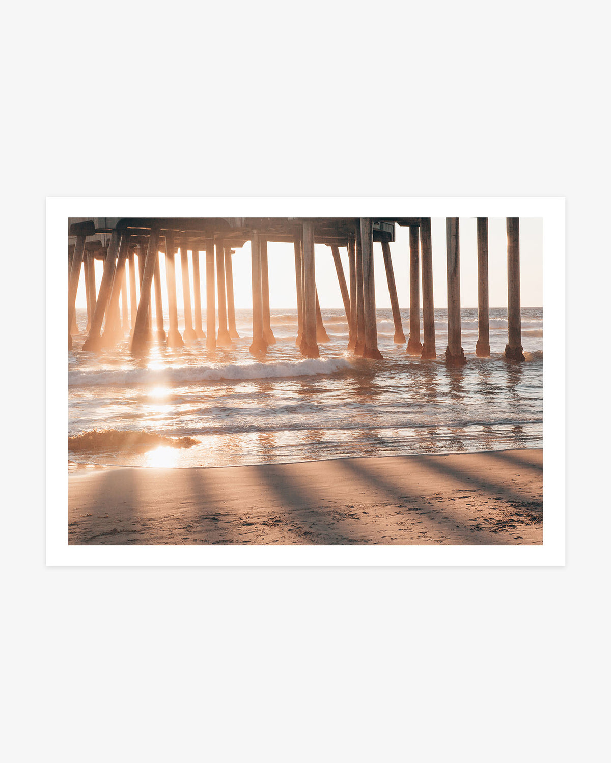 Huntington Beach Pier wall art featuring golden sunlight streaming between the wooden pilings and reflecting across the Pacific waves and wet sand.