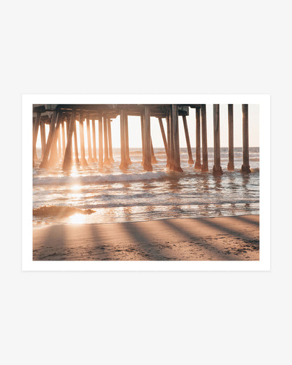 Huntington Beach Pier wall art featuring golden sunlight streaming between the wooden pilings and reflecting across the Pacific waves and wet sand.