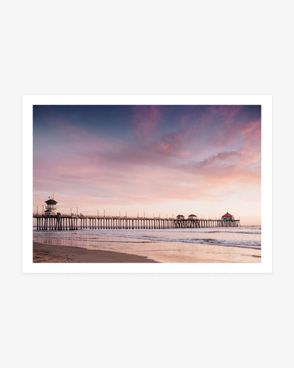 A framed art print featuring a scenic photograph of a pier extending over a beach under a pastel pink, purple, and blue sunset sky.