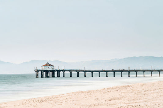 Manhattan Beach Pier photography featuring the iconic roundhouse over calm Pacific Ocean waters, with sandy shoreline and hazy coastal mountains in soft blue and beige tones.